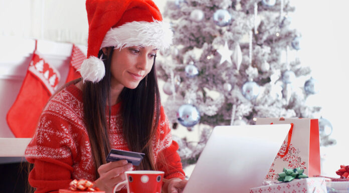 Młoda kobieta w świątecznym swetrze i czapce Mikołaja siedzi przy komputerze i trzyma w ręce kartę płatniczą.